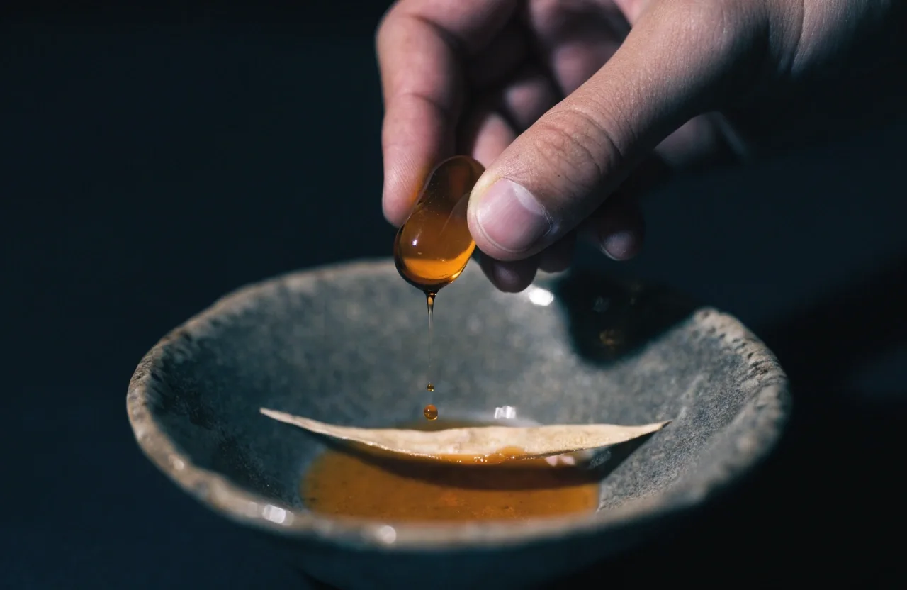 Macro of an opened softgel with amber content pooling beside a dried botanical strip on a dark matte surface